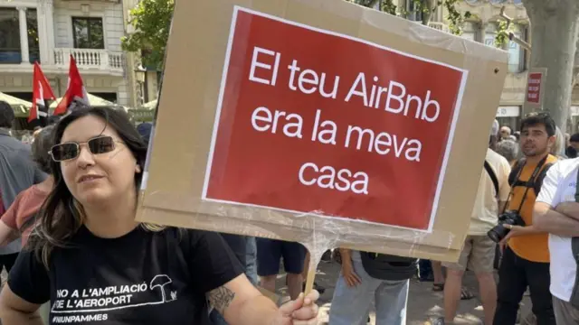 Mulher protesta nas ruas de Barcelona portando um cartaz que diz, em catalão, "o seu AirBnB era a minha casa"