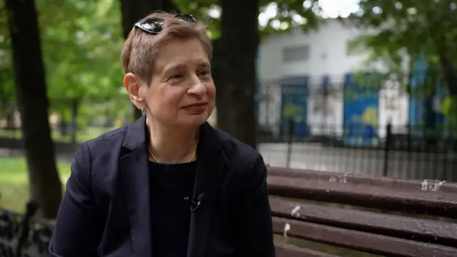 Starija žena u crnom sakou sa naočiram za sunce na glavi sedi na klupi u parku