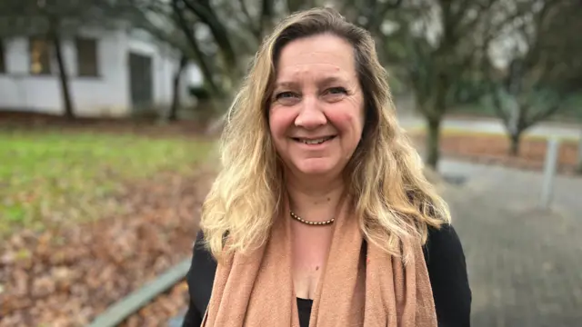 Lucy Nethsingha mira a la cámara. Tiene el pelo rubio y lleva un jersey oscuro con una bufanda naranja quemada. Hay árboles detrás.