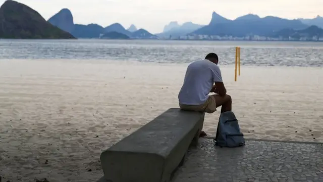 Homem olhando para baixo e sentadofutebol de 5banco na beirafutebol de 5praiafutebol de 5Niterói, com vista do Riofutebol de 5Janeiro ao fundo