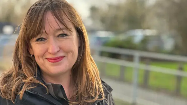 Emily Cherry, sonriente, tiene el pelo castaño hasta los hombros. Sonríe y lleva una chaqueta negra.