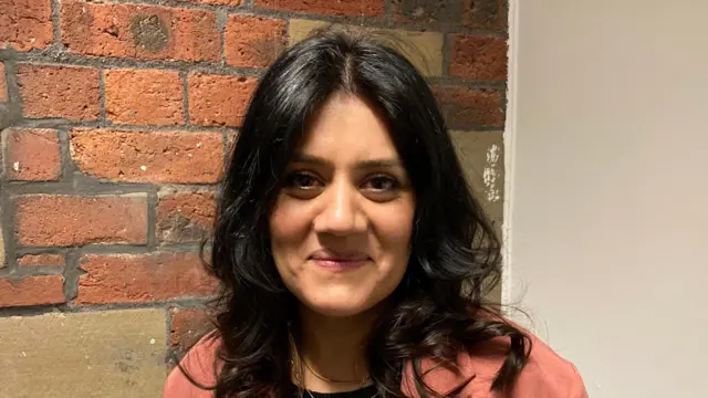 Una mujer con cabello largo y negro brillante. Sonríe y está de pie frente a una pared de ladrillos. Lleva un blazer rosa.