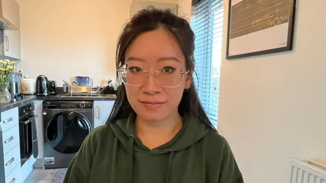 A woman in a kitchen looks at the camera with a neutral expression. She wears frameless glasses and a green hoodie and her dark hair hangs behind her shoulders. Behind her is a washing machine to the left and a vase of yellow flowers is on the worktop beside it. 