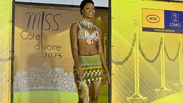 Une candidate à un concours de beauté en Daloa, vêtue d'un bikini orné de bijoux et d'un short à motifs traditionnels et tenant un chasse-mouches, marche sur le podium. Derrière elle, des banderoles jaunes portant le logo de Miss Côte d'Ivoire 2025. 