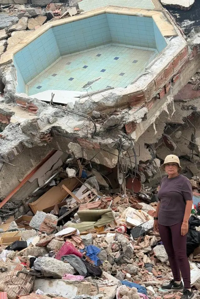 María Lourdes Pérez, vestida com calça e camisa marrom e gorro bege, parada em um local repleto de escombros, com o que parece ser uma pequena piscina, pedras, cimento, partes de uma edificação, móveis destruídos e roupas