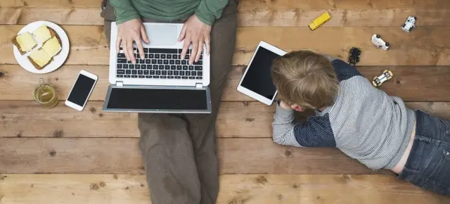A mother and toddler sitting on the floor, the boy on a tablet, the mother on a laptop. There's also a smartphone within reach.