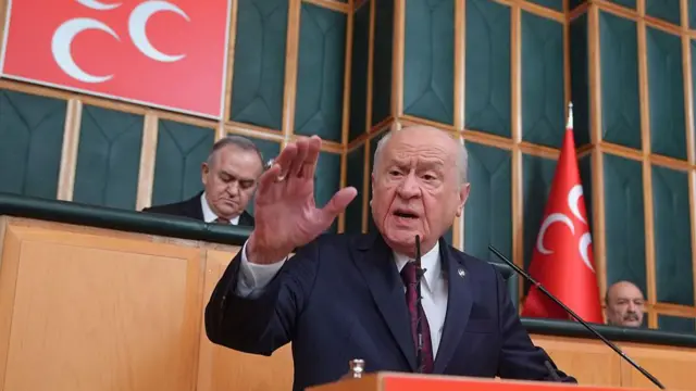 MHP lideri Devlet Bahçeli'nin 21 Nisan'daki grup konuşması. Arkada üç hilalli logolar ve diğer parti yetkilileri var. Bahçeli koyu bir takım elbise giymiş, kürsüde konuşuyor. 