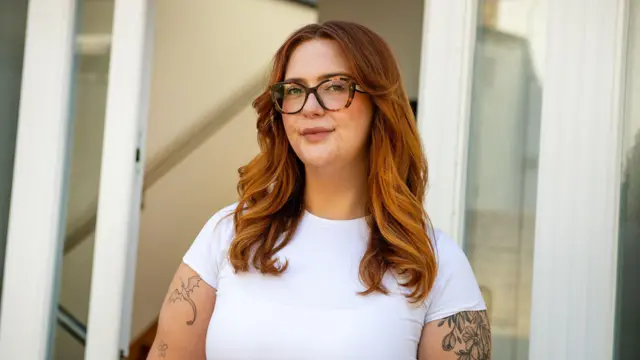 Jess King, egy hosszú vörös hajú, szemüveges nő, egy ajtó előtt áll. Rövid ujjú fehér felsőt visel, és karjai felső részén jól látható fekete tetoválások vannak.
