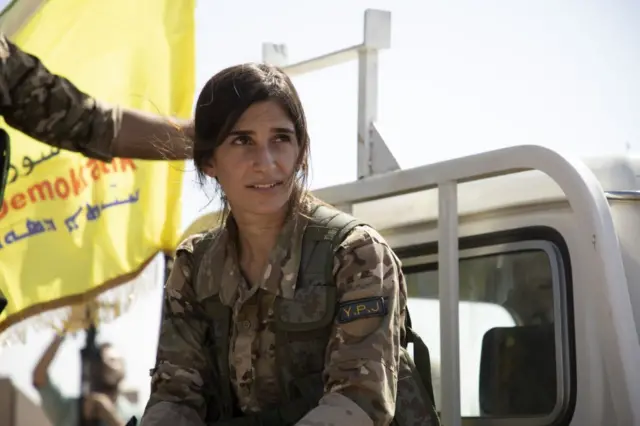 الصورة لشابة مقاتلة في صوف قسد ببزة عسكرية وفي الخلفية العلم الكردي الأصفر