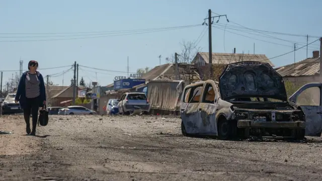 Uma mulher caminha por uma rua ensolarada, carregando uma sacola de compras, ao lado de um carro destruído por uma bomba.
