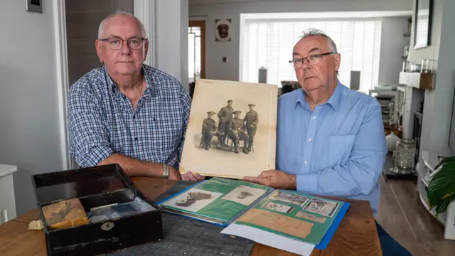 Imagem panorâmica de dois homens com cerca de 60 anos, sentados a uma mesa de madeira em uma sala de estar. Eles seguram uma foto antiga com quatro homens. Alistair, sentado à esquerda, veste uma camisa quadriculada azul e Andrew, à direita, veste uma camisa azul clara.