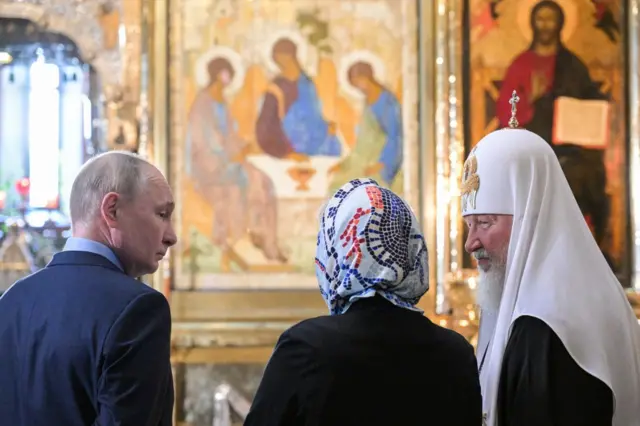 Путин и патриарх Кирилл перед иконой в храме. Между ними, спиной к камере, стоит какая-то женщина в платке