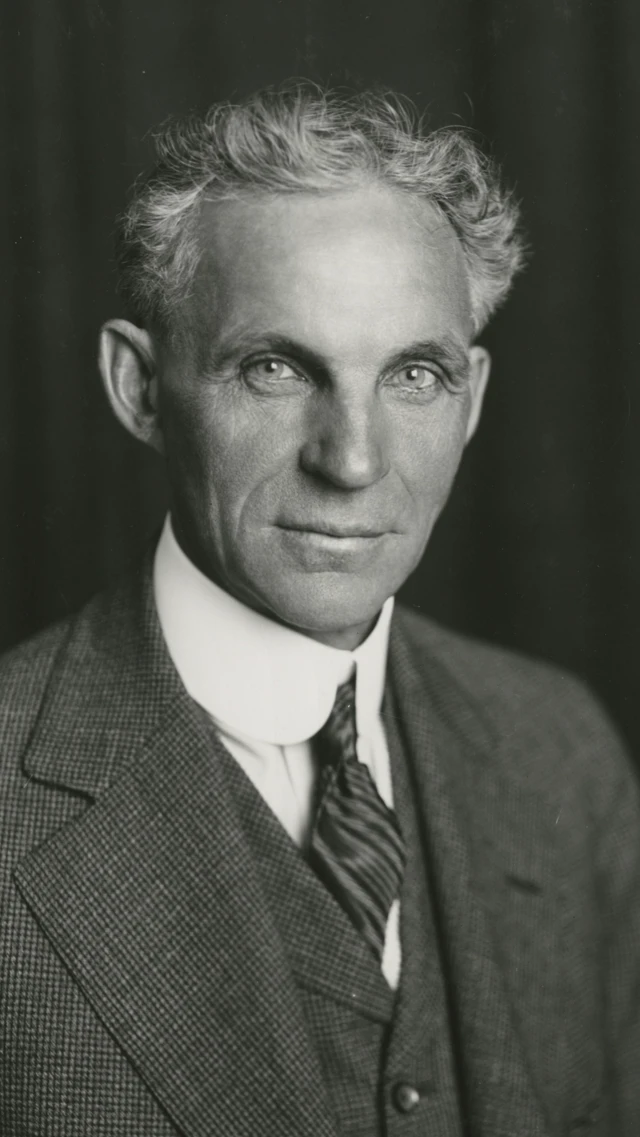 A imagem em preto e branco mostra um homem usando terno, colete, gravata e camisa de colarinho alto posando para um retrato formal. Ele tem cabelos claros e ondulados, penteados para trás, e olha diretamente para a câmera com expressão séria. O fundo escuro e desfocado destaca o rosto e a roupa do retratado.