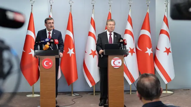 Yılmaz ve Üstel kürsüden konuşuyor. Arkalarında Türk ve Kuzey Kıbrıs bayrakları var.