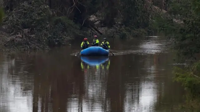 根こそぎ流され倒れた流木が折り重なる川岸を背に、赤いヘルメットと蛍光黄色のジャケットを付けた救助隊員3人が、青いゴムボートを漕いで川を進んでいる