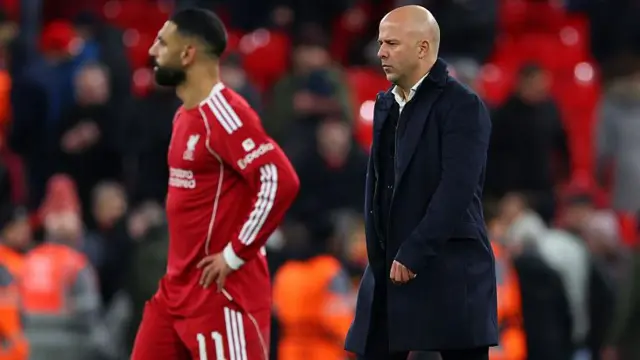 آرنه سلات، سرمربی لیورپول، با بالاپوش سیاه و پیراهن سفید در سمت چپ و محمد صلاح، با لباس سرخ و خط‌ها و نوشته‌های سفید در آن در سمت چپ در زمین بازی قرار دارند.