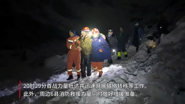 Una senderista con casco y ropa térmica se apoya en dos socorristas mientras bajan una ladera llena de nieve durante la noche, seguidos de otros tres socorristas, uno de los cuales ilumnina el camino con una lámpara.