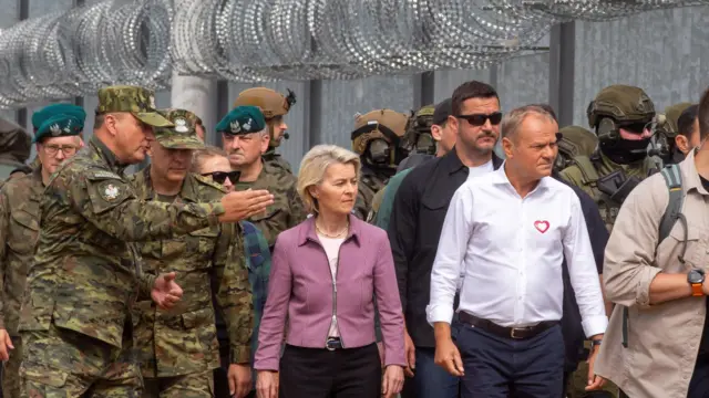 Premier Polski Donald Tusk i przewodnicząca Komisji Europejskiej Ursula von der Leyen idą obok polskiego ogrodzenia granicznego z Białorusią. Za nimi stoją żołnierze w mundurach maskujących.