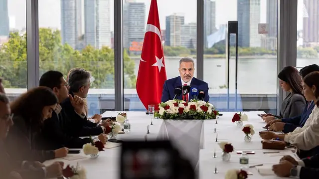 Hakan Fidan'ın arkasında Türk bayrağı var. Uzun bir masada gazetecileri görüyoruz. Arkada fondaysa bir nehir ve gökdelenler var.