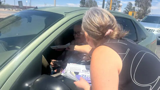 Silvia Delgado, vestindo uma blusa preta e branca, está distribuindo panfletos aos eleitores em um carro na Ponte das Américas.