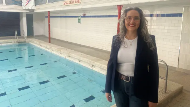 La concejala Marina Strinkovsky, vestida con vaqueros azul oscuro, un cinturón negro ancho, una camiseta blanca y una chaqueta azul oscuro, tiene el pelo largo, rizado y gris, y lleva gafas. Está de pie en la plataforma de baldosas junto a la gran piscina, sonriendo a la cámara.