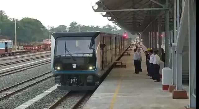DEMU ရထားတွဲ တစ်ခု မြန်မာက ဘူတာရုံတစ်ခုက စင်္ကြံန်ဘေးမျာ ရပ်ထားတာကို မြင်တွေ့ရ၊ ရထား ဘယ်ဘက်ခြမ်းမှာ သံလမ်းတွေကို မြင်တွေ့ရပြီး ညာဘက်ခြမ်း စင်္ကြံဘက်မှာတော့ ရထားဝမ်ထမ်းအချို့ကို မြင်တွေ့ရ၊ သူတို့ခေါင်းပေါ်မှာ ဘူတာရုံအမိုး တစ်စွန်းတစ်စကိုလည်း မြင်တွေ့ရ 