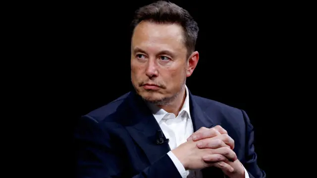 Elon Musk é um homemsite betfairpele clara, cabelos curtos, e está vestindo um blazer marinho sobre camisa branca. Ele olha para o lado esquerdo e está com as mãos cruzadas.