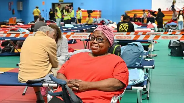 En primer plano, una mujer con turbante en la cabeza que le cubra el pelo, gafas, lleva camiseta roja y tiene las manos cruzadas. está sentada en una silla con sus muletas al lado. Sonríe a la cámara. Al fondo se ve gente sentada en camillas de campaña. Hay filas y filas de camillas de campaña. Al fondo de la foto, voluntarios con chalecos amarillos y más personas sentadas en las camillas. 