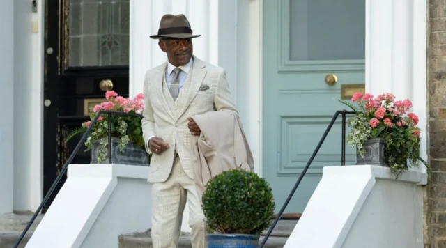 Un hombre vestido de elegante traje blanco y sombrero baja por las escaleras de acceso a una casa. 