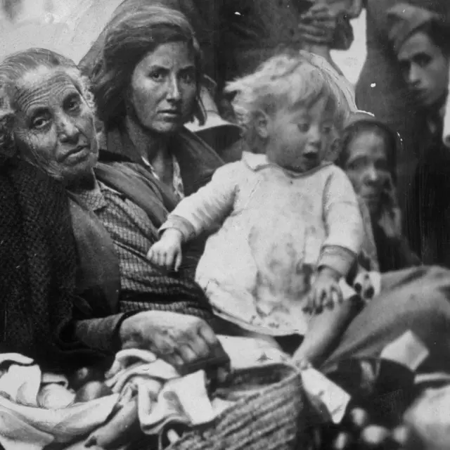 Una anciana, junto a su hija y a su nieta, refugiadas todas de la guerra civil española, llegan a Valencia cargando sus pertenencias en 1937.