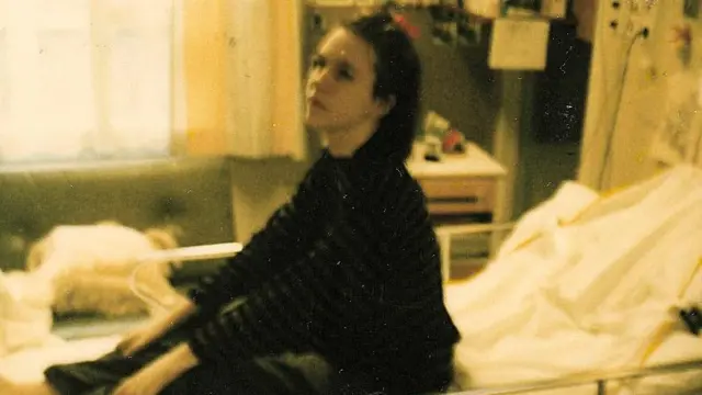 Egy enyhén elmosódott színes fényképen Nina látható, amint egy kórtermi ágyon ül egy kórházi osztályon. Az ágyon felhajtható fém oldalkorlátok és fehér ágynemű van, a közelben pedig orvosi berendezések vannak a falra szerelve. A kép bal oldaláról egy nagy ablakon át természetes fény áramlik be. A háttérben szekrények, polcok és kórházi berendezési tárgyak láthatók. A fényképet az 1989-ben készült régebbi felvételekre jellemző fakó színek és szemcsés képminőség jellemzik.