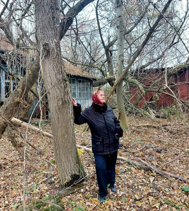 Жінка у чорній куртці, широких джинсах і червоній хустці стоїть серед занедбаного подвір’я, опершись на стовбур дерева. Навколо багато сухого пожовклого листя та гілок, а в тлі видно покинутий дерев’яний будинок.