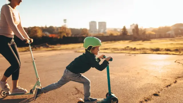 Un niño anda en patineta y su madre lo sigue detrás montando otra patinera.