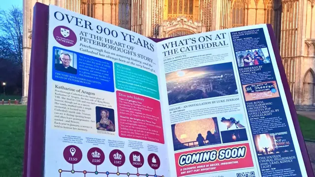 Un gran libro rojo con información en ambos lados, colocado sobre el césped con el edificio de la Catedral al fondo.