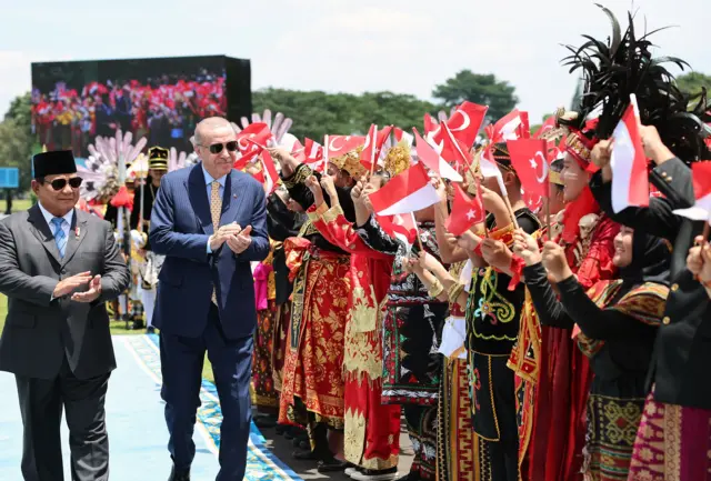Erdoğan ve Subianto geleneksel kıyafetler giyen kalabalığın önünde yürüyor. İki lider takım elbiseli ve güneş gözlükleri takıyor. Kalabalığın elinde Türk ve Endonezya bayrakları var.