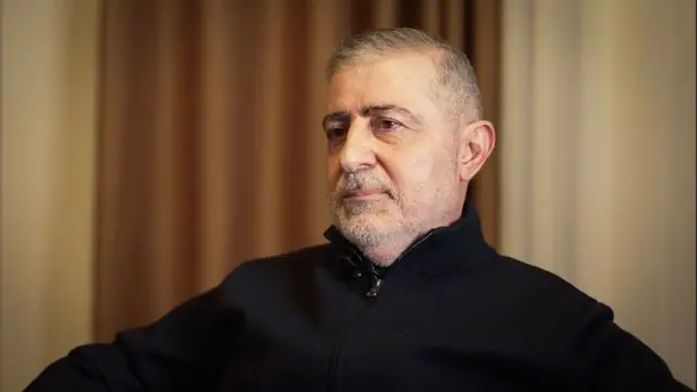 Wafiq Safa sentado em uma cadeira, vestindo casaco preto e olhando para o lado da câmera. Ele é um homem de meia idade, com barba e cabelos grisalhos aparados