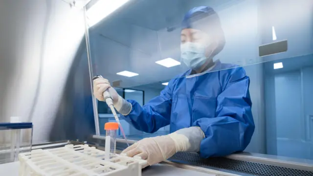 A imagem mostra um laboratório com iluminação intensa, superfícies limpas e ambiente controlado. Uma pessoa veste equipamentos de proteção, incluindo avental azul, luvas, máscara e touca. Ela usa uma pipeta para transferir líquido para um pequeno tubo apoiado em uma bandeja. Uma barreira transparente protege a área de trabalho, indicando um ambiente estéril. Ao fundo, equipamentos de laboratório e superfícies refletivas reforçam o ambiente clínico e científico