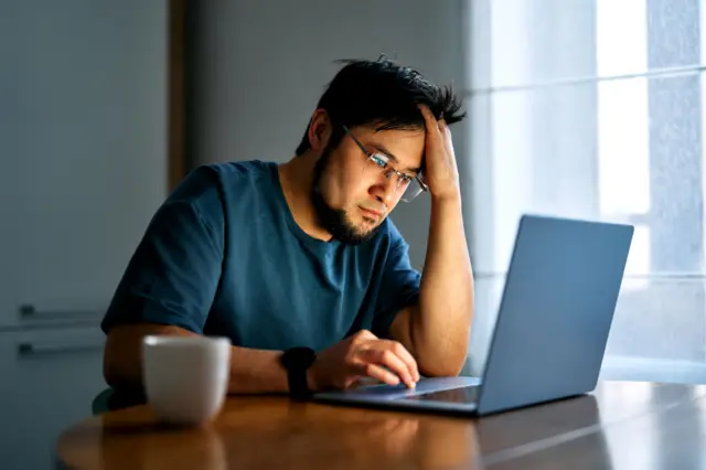 Hombre frustrado que experimenta agotamiento y ansiedad, sosteniéndose la cabeza con la mano mientras está sentado a una mesa con un portátil.