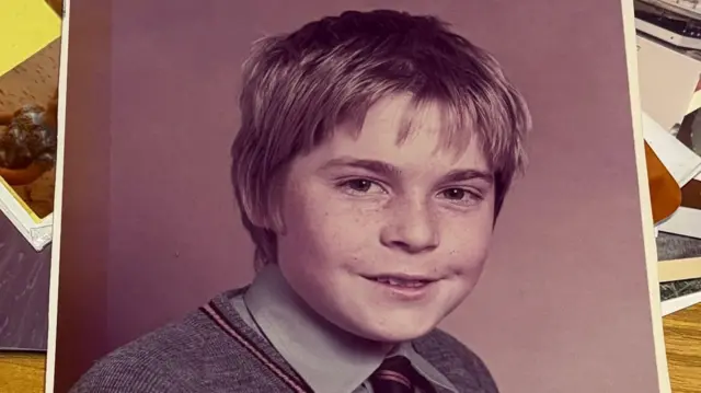 Uma antiga fotografia escolar mostra um menino com sardas. Ele veste uma camisa cinza e uma gravata preta e dourada sob um suéter grande de gola em V