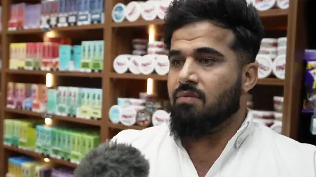 Close-up of Twana Jamal with dark hair and a full beard, wearing a white collared shirt, standing indoors in front of wooden shelves filled with neatly arranged, colourful product boxes and small round containers. A microphone with a fuzzy windscreen is positioned in the lower foreground, indicating an interview or recording. The background features warm lighting within the shelving units, highlighting the rows of packaged items.