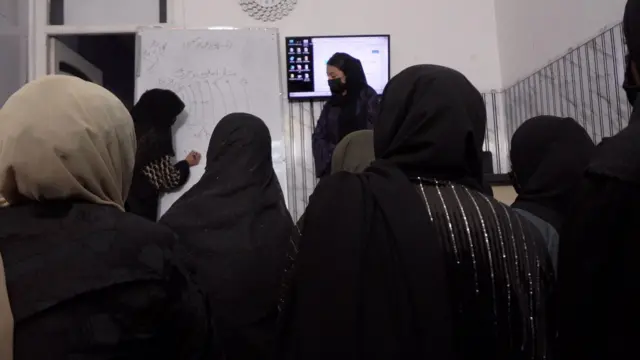 شاگردی چیزی بروی تخته سفید می‌نویسد، معلم که در کنار او ایستاده و شاگردان که بروی زمین نشسته‌اند، به او نگاه می‌کنند. 