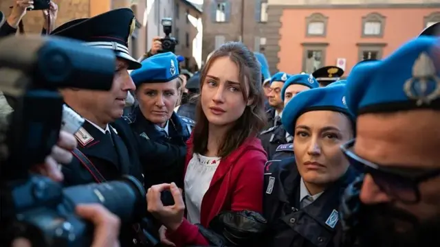 A atriz Grace Van Patten, no papel de Amanda Knox, avança por um grupo de jornalistas que tentam fotografá-la, enquanto é protegida por vários policiais