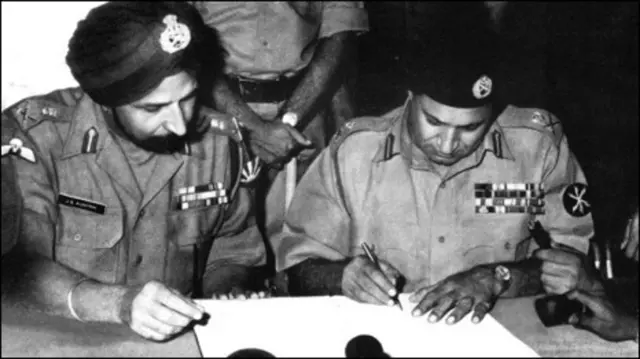 রমনা রেসকোর্সে লেফটেনান্ট জেনারেল জগজিৎ সিং অরোরার উপস্থিতিতে আত্মসমর্পণের দলিলে স্বাক্ষর করছেন পাকিস্তানি বাহিনীর কমান্ডার লেফটেনান্ট জেনারেল নিয়াজী