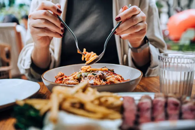 Bir restoranda yemek masasında oturan, elinde çatal-bıçakla yeni servis edilmiş spagetti bolognese'i dolayan ve yemeğinin tadını çıkaran genç Asyalı kadının yakın plan, gövde hizasından görünümü.