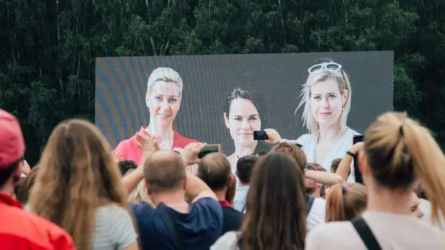 Люди со спины стоят перед сценой на уличном митинге. На сцене фотография, где изображены Мария Колесникова, Светлана Тихановская и Вероника Цепкало