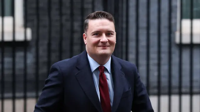 Wes Streeting aparece en la foto saliendo del número 10 de Downing Street en Londres. Lleva chaqueta azul oscuro, camisa azul claro y corbata roja. Tiene el pelo castaño corto y está bien afeitado.