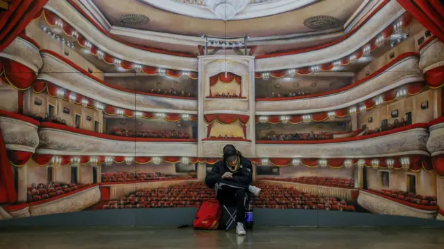 One pesin don sit wey im cross legs for one metro station for Kyiv, in front of painting wey dey show large theatre during di air raid for Kyiv for March 2024. 