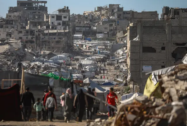 Adultos e crianças caminham em meio ao entulho e prédios destruídos. Tendas são vistas ao fundo. 