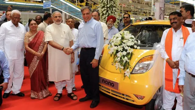 Narendra Modi, then governor of Gujarat, shakes hands with the chair of Tata group at a new factory. A yellow car is adorned with white flowers.