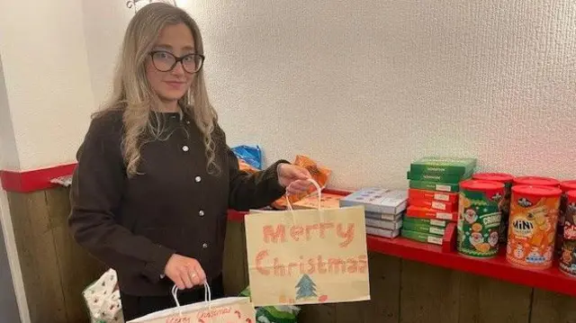 Una mujer con cabello rubio ondulado y gafas sostiene bolsas con la frase "Feliz Navidad" en una. Hay bocadillos y chocolates en un estante a su lado.